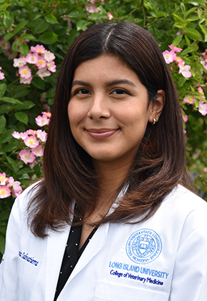 Dr. Genesis Salvatierra with a soft smile in front of a bush blooming with small pink flowers. She has on a black polka dot shirt and her whitecoat from Long Island University.