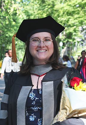 Dr. Chloe Kenney smiling outside on a university campus. She has on her doctoral robe, stole and cap, round glasses, and is holding a bouquet of flowers.