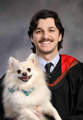 Dr. Christopher Kouris sitting for a professional portrait in front of a grey, heathered backdrop. He is smiling while wearing his black doctoral robe and red hood. He is holding a white Pomeranian who has on a blue bandanna featuring yellow rubber ducks and is looking to be smiling as well.