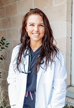 Dr. Jessica "JJ" Barber smiling outside in front of a brick wall. She has on navy scrubs, her BluePearl whitecoat and has a stethoscope around her neck.