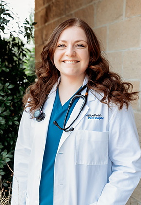 Dr. Brianna Huff smiling outside in front of a bush next to a brick building. She is wearing her BluePearl whitecoat, blue scrubs and has a stethoscope around her neck.