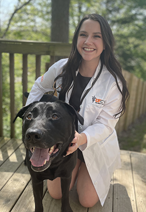 Dr. Sierra Johnson smiles while sitting on her knees outdoors on a deck. Her black lab is next to her, seemingly smiling. Dr. Johnson is wearing a black dress and her whitecoat.
