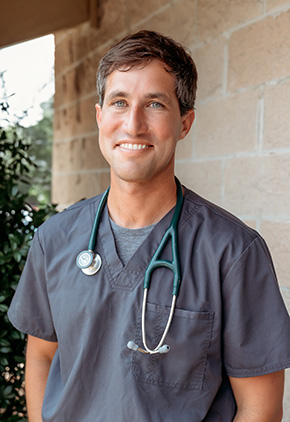 Dr. Justin Loudermilk smiling outside in front of a bush next to a brick building. He has on grey scrubs with a dark green stethoscope around his neck.