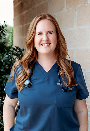Dr. Ashley Schug smiling outside in front of a bush next to a brick building. She has on navy blue scrubs with a stethoscope around her neck.