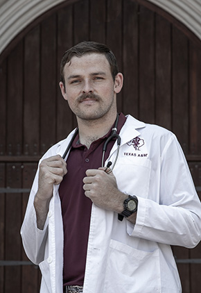 Dr. Kadden Kothmann standing strong in front of an older building's wood door. He has on a maroon shirt and his Texas A&M whitecoat with his arms bent 90 degrees and his hands on either end of his stethoscope.