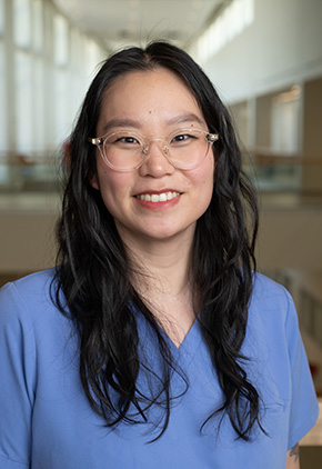 Dr. Jenna Cao smiling in an academic building. She has on clear, circular glasses and light blue scrubs.