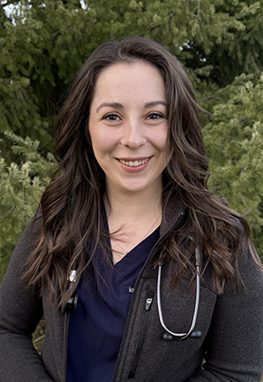 Dr. Meghan Feeney smiling outside in front of pine trees. She has on navy blue scrubs, a heather zip-up and a stethoscope around her neck.