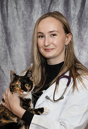 Dr. Gwendolyn Hall smiling for a professional portrait in front of a grey backdrop. She has on a black top, her whitecoat and has a stethoscope around her neck. She is holding her black and orange mixed cat.
