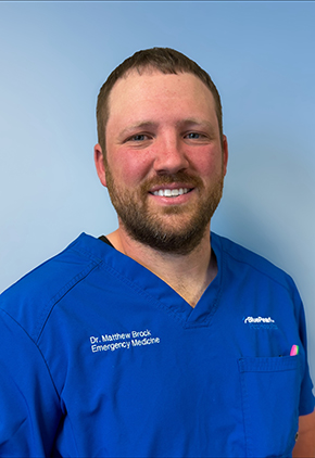 Dr. Matthew Brock smiles for a headshot in front of a light blue wall. He is wearing royal blue scrubs.