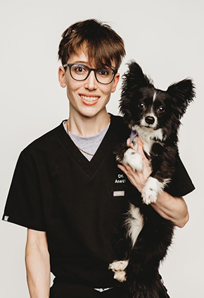 Dr. Erika Militana, wearing black scrubs and glasses, smiles while holding her black and white Chihuahua mix, Tuxedo, against a plain background.