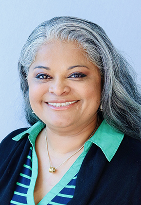 Dr. Aarti Wiliams smiling for a professional photo in front of a white wall. She has on a navy cardigan and green and navy polo.