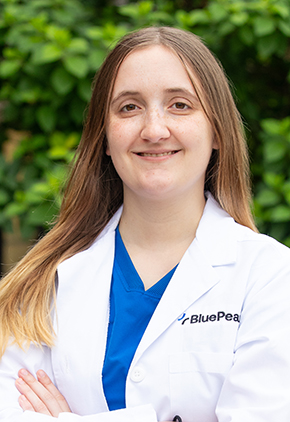 Dr. Megan Smith smiling outside in front of a lush, green tree. She has on royal blue scrubs and her BluePearl whitecoat.