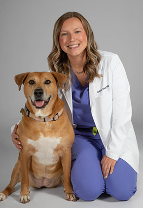 Dr. Madison Colon smiles for a professional studio photo while kneeling next to her bully terrier mix. She is wearing powder blue scrubs and her BluePearl whitecoat.