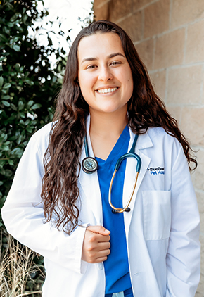 Dr. Cassidy Czaykowsky smiling outside in front of a bush next to a brick wall. She has on royal blue scrubs and her BluePearl white coat with a teal stethoscope around her neck.