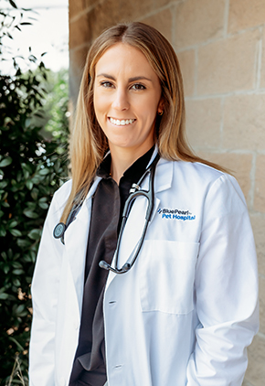 Dr. Kayla Schonvisky smiling outside in front of a bush next to a brick wall. She is wearing black scrubs, her BluePearl white coat and has a stethoscope around her neck.