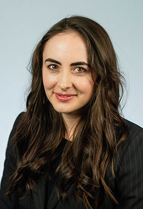 Dr. Lauren Ryon smiles softly for a headshot in front of a light blue backdrop. She is wearing a black suit jacket and black dress shirt.