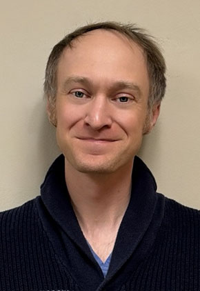 Dr. Matthew Pickens smiles for a professional headshot in front of a beige background. He is wearing a dark navy sweater over blue scrubs.
