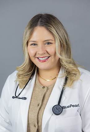 Dr. Kristyn Cunningham smiles for a professional headshot in front of a grey background. She is wearing a tan, buttoned-up cardigan, her BluePearl white coat and has her stethoscope around her neck.