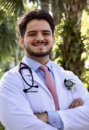 Dr. Robert Gonzalez smiling outside in front of trees. He is wearing a light blue dress shirt, dusty red tie and his BluePearl whitecoat with a stethoscope around his neck.