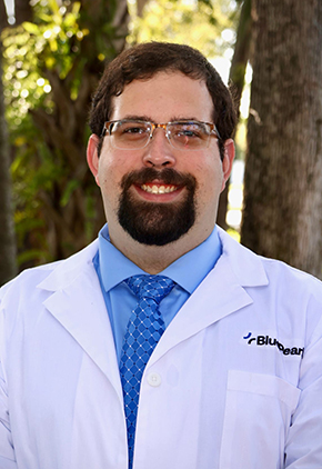 Dr. Joshua Bard smiling outside in front of trees. He is wearing a blue dress shirt, blue tie and his BluePearl whitecoat.