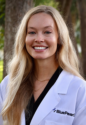 Dr. Cardinal Hastings smiles outside in front of tropical trees. She has on a black top and her BluePearl whitecoat.