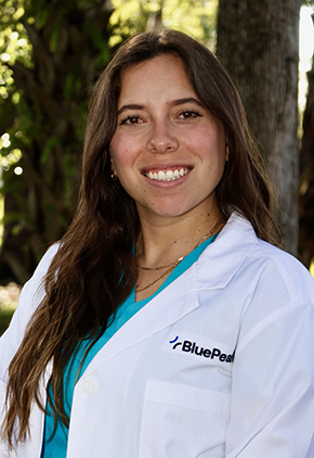 Dr. Alexandra López De Victoria smiling outside in front of trees. She is wearing a sea blue top and her BluePearl whitecoat.