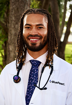 Dr. McCoy Williams smiling outside in front of tropical trees. He is wearing a dress shirt and blue tie with his BluePearl whitecoat, and a stethoscope is around his neck.