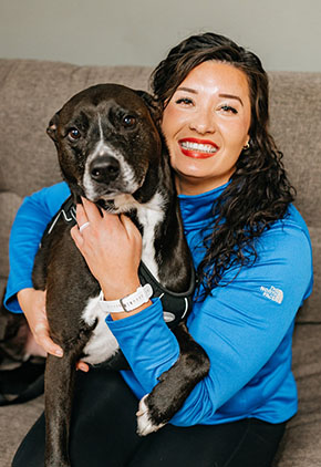 Kayla Wilcox smiles while sitting on a tan couch next to her black and white pit bull, Drake. She is wearing a light blue North Face quarter-zip and hugging Drake.