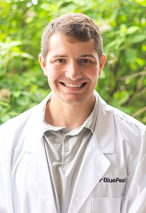 Dr. Grant Hoeger smiles outside in front of a leafy, green tree. He is wearing a grey polo and his BluePearl whitecoat.