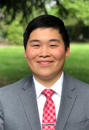Dr. Ryan Chang smiles in a large park clearing. He is wearing a grey suit, white dress shirt and red & grey argyle tie with a silver tie clip.