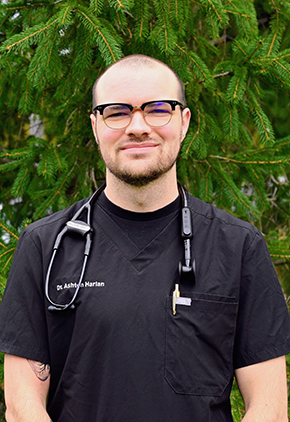 Dr. Ashton Harlan smiles outside in front of an evergreen pine. He is wearing a black shirt and has his stethoscope around his neck.