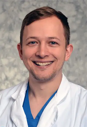 Dr. Christopher Kuechle smiles for a professional studio headshot in front of a marble grey background. He is wearing blue scrubs and his whitecoat.