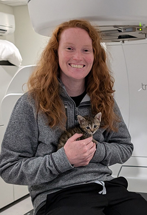 Dr. Kelsey Duggan smiling while holding a kitten and sitting on an exam table in an imaging room. She is wearing black pants and a grey longsleeve.