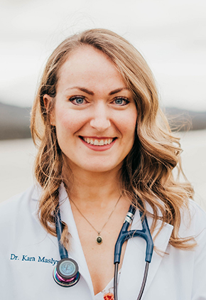 Dr. Kara Maslyn smiles outside on the shores of a lake. She is wearing a white floral shirt, her whitecoat and has a stethoscope around her neck.