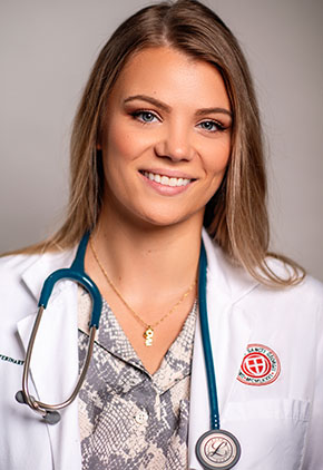 Dr. Adriana Kalaska smiles in a professional headshot taken in front of a gray wall. She wears a snake-patterned shirt under a St. George’s University white coat, with a navy stethoscope around her neck.