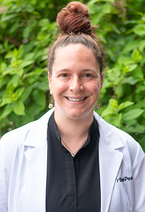 Dr. Sarah Kowarski smiles outside in front of a green, leafy tree. She is wearing a black dress shirt and her BluePearl whitecoat.
