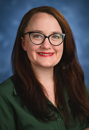 Dr. Erin Perry smiles for a professional headshot in front of a classic blue backdrop. She is wearing a forest green dress shirt with matching green glasses.