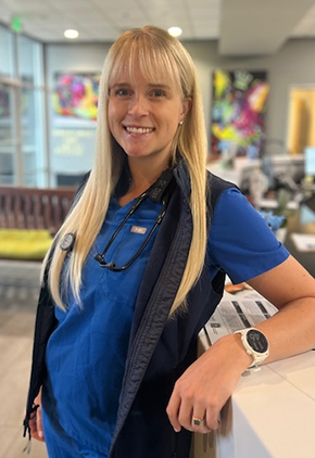 Dr. Leanne Diss smiles in the lobby of our Reno hospital. She is wearing blue scrubs, a black vest, and has a stethoscope around her neck while leaning against the front desk.