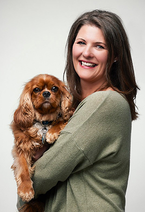Dr. Leah Gibbs smiles for a professional photo. She is wearing a green long-sleeved shirt and is holding her dog in her arms.