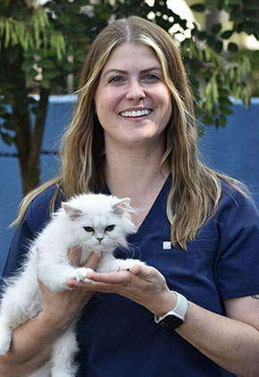 Dr. Megan Korpita smiles outside while holding a white kitten in her rams. She is wearing navy scrubs and a smart watch.