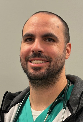 Dr. Christopher Presby smiles for a headshot in the hospital. He is wearing teal scrubs, a grey zip-up hoodie and has a stethoscope around his neck.