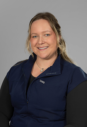 Dr. Heather Turnbull smiles for a professional headshot in front of a grey studio background. she is wearing a black blouse and a navy vest.