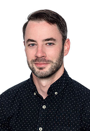 Dr. Michael Hale smiles in front of a white studio backdrop for a professional headshot. He has on a button up shirt that is black with very small white polka dots.