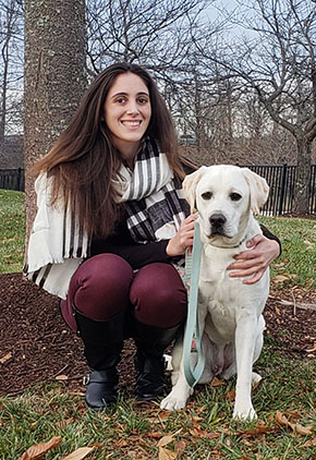 Dr. Rebecca Weiske smiles outside on a fall day. She is squatting in front of a tree and next to her yellow lab.