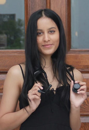 Dr. Nikki Shinde smiles on the steps in front of a university building. She has a black tank top on and is holding on to either end of her stethoscope around her neck.