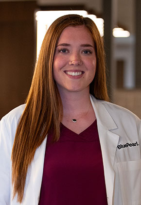 Dr. Julia Honan smiles for a headshot in the lobby of BluePearl Pet Hospital. She is wearing a maroon blouse and her whitecoat.