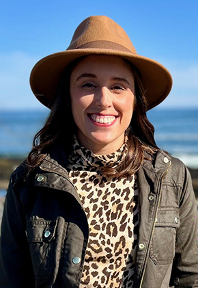 Dr. Allison Wilson smiles outside with a large body of water in the background. She is wearing an army green jacket, leopard turtleneck and a brown hat with stiff, all-around brim.