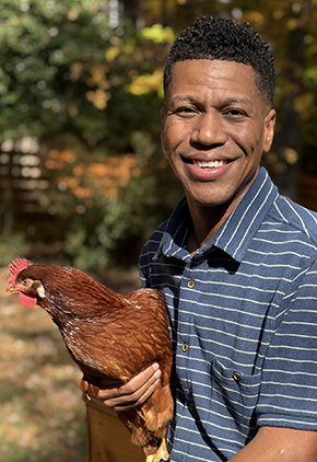 Dr. Raphael Malbrue smiles outside while holding a rooster. He is wearing a blue shirt with small white lines.