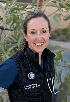 Dr. Catriona Forrest smiles outside in front of a leafy tree. She wears a navy scrub top, a black BluePearl vest and has a stethoscope around her neck.