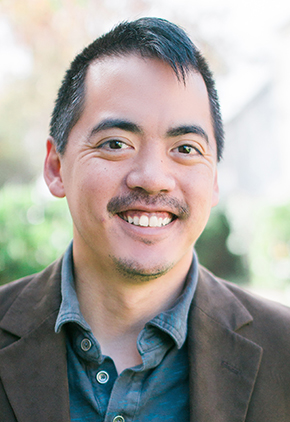 Sam Tanng smiles outside for a professional headshot. He is wearing a tweed suit jacket and a denim blue button-up.